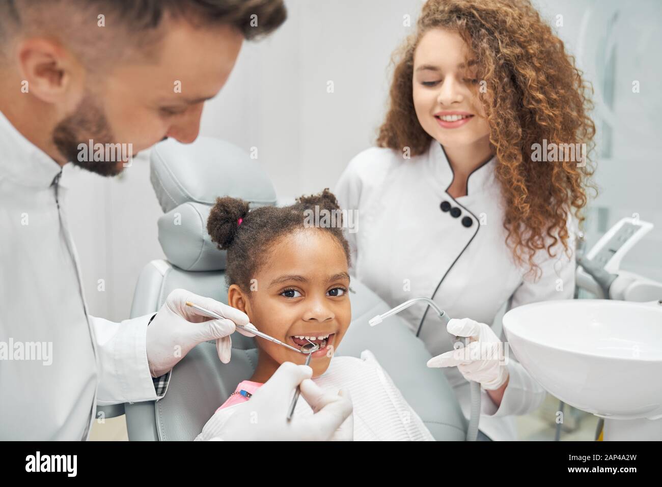 Docteur En Gants Médicaux Noirs Examine Les Dents De La Jeune Fille En  Utilisant Un Microscope Dentaire Et Un Miroir Dans Le Bureau Du Dentiste.  La Femme A Un Bavoir Du Patient
