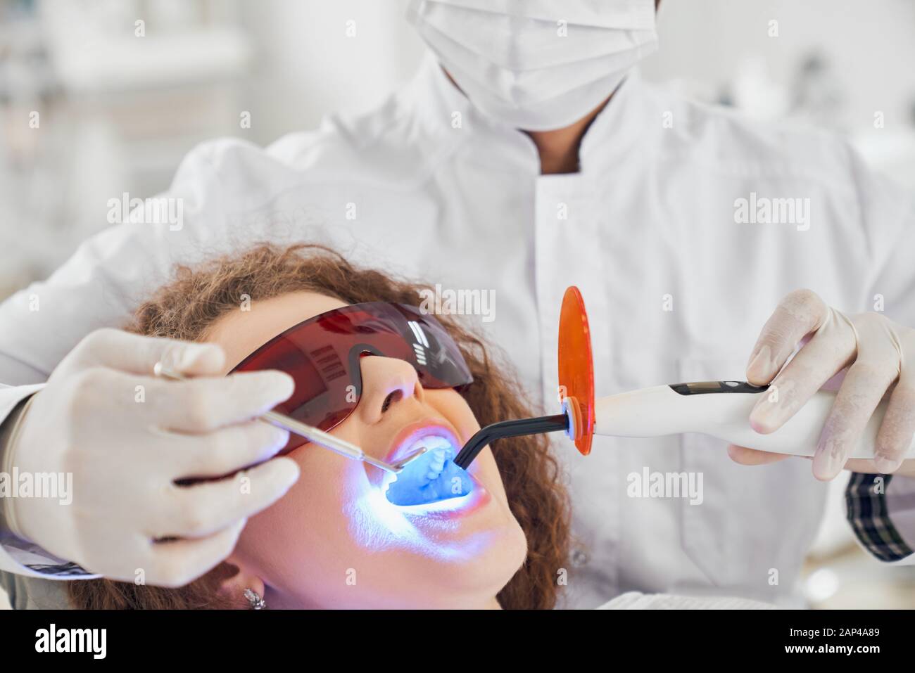 Jolie dame avec des cheveux couchés dans une chaise dentaire dans des lunettes de protection à bouche ouverte tandis que médecin mâle dans des verres blancs faisant la procédure de blanchiment. Concept de soins dentaires Banque D'Images