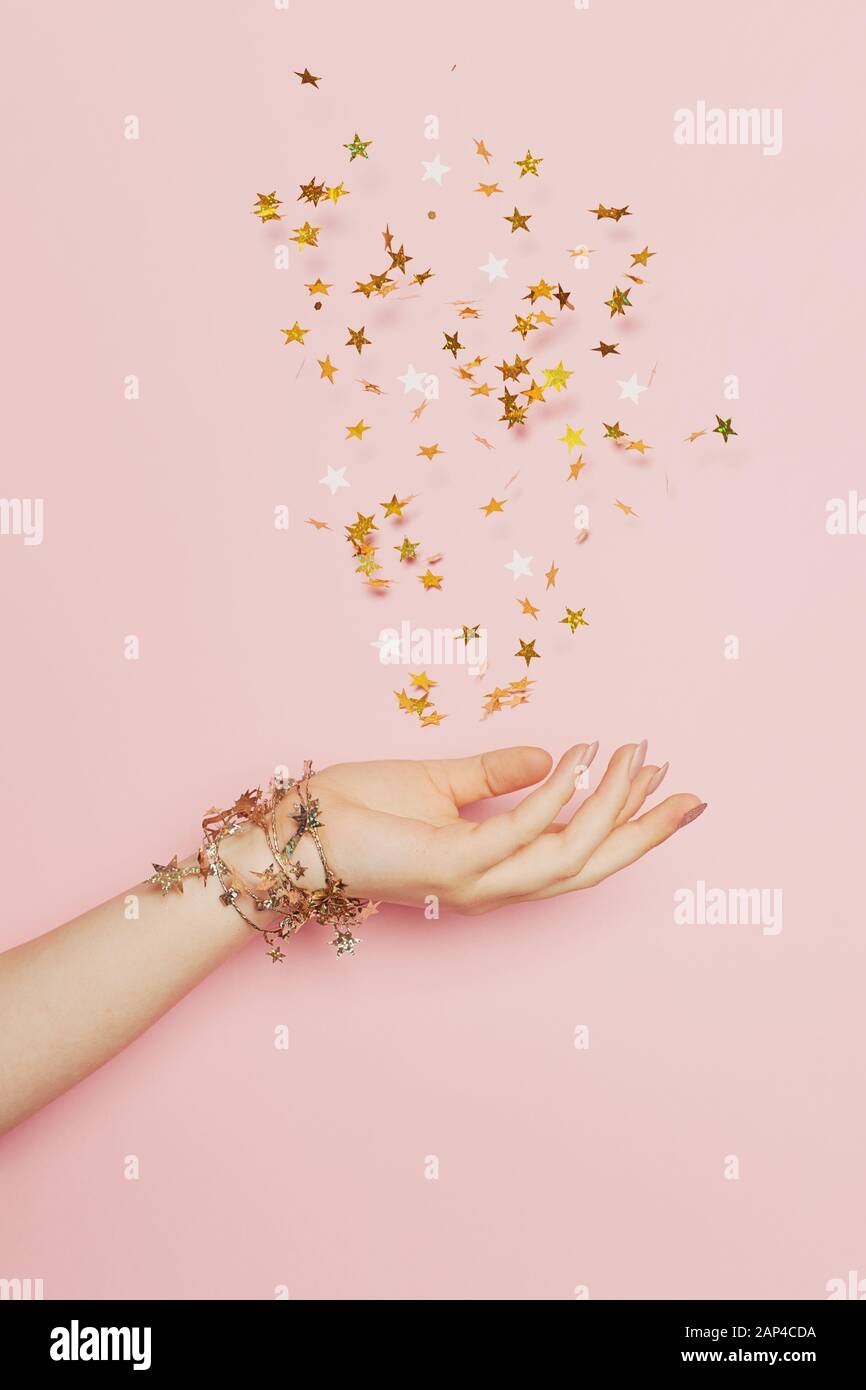 Concept de fête, mariage et fête d'anniversaire. Les étoiles confettis d'or se gonflent dans la main féminine sur fond rose pastel Banque D'Images