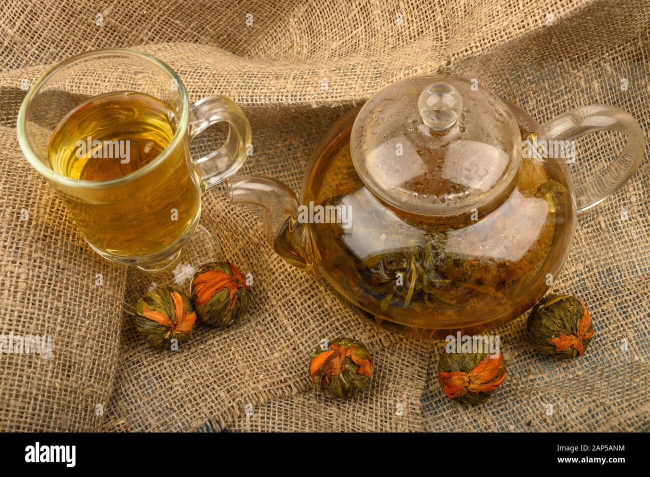 Thé aux fleurs préparé dans une théière en verre, un verre de thé et des boules de thé aux fleurs sur fond de tissu à l'Homespun rugueux. Gros plan Banque D'Images