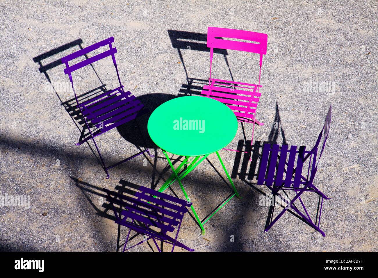 Vue en grand angle sur une table ronde verte avec quatre chaises pliantes en bois rose et violet en plein air en plein soleil jettent des ombres sur un sol gris en gravier - Pr Banque D'Images