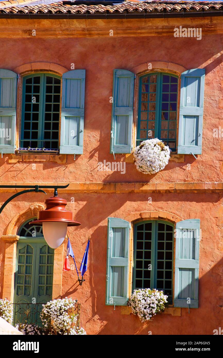 Vue sur la façade lumineuse méditerranéenne de la maison d'ocre rouge avec volets bleus, portes vertes, pots de fleurs - Roussillon en Provence, France Banque D'Images