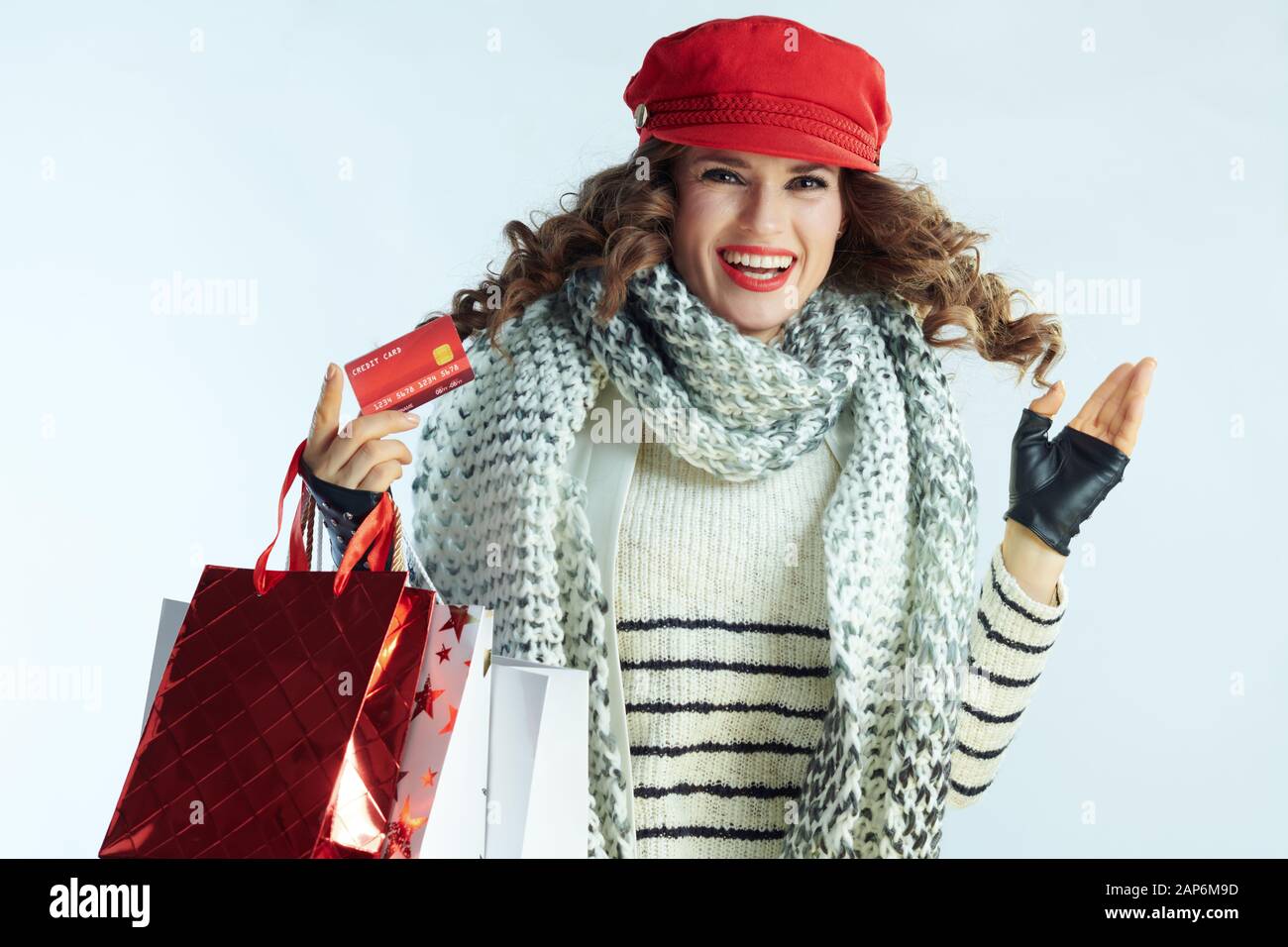 Élégant sourire 40 ans femme aux longs cheveux de brunette en pull, écharpe  et red hat with shopping bags et saut de carte de crédit sur la lumière  d'hiver bl Photo Stock -