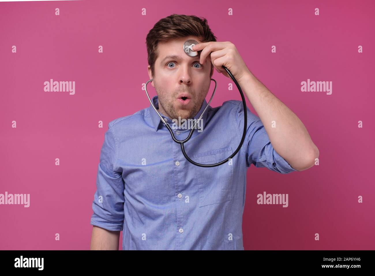 Jeune homme avec stéthoscope écoutant ses battements de cœur sur la tête. Le gars stupide ne sait pas comment utiliser l'outil de médecine. Studio tourné Banque D'Images