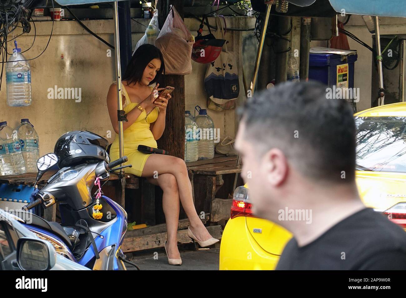 Bangkok, Thaïlande - 26 décembre 2019 : femme transgenre, ladyboy, assise sur banc et par téléphone. Homme au premier plan la regardant. Banque D'Images
