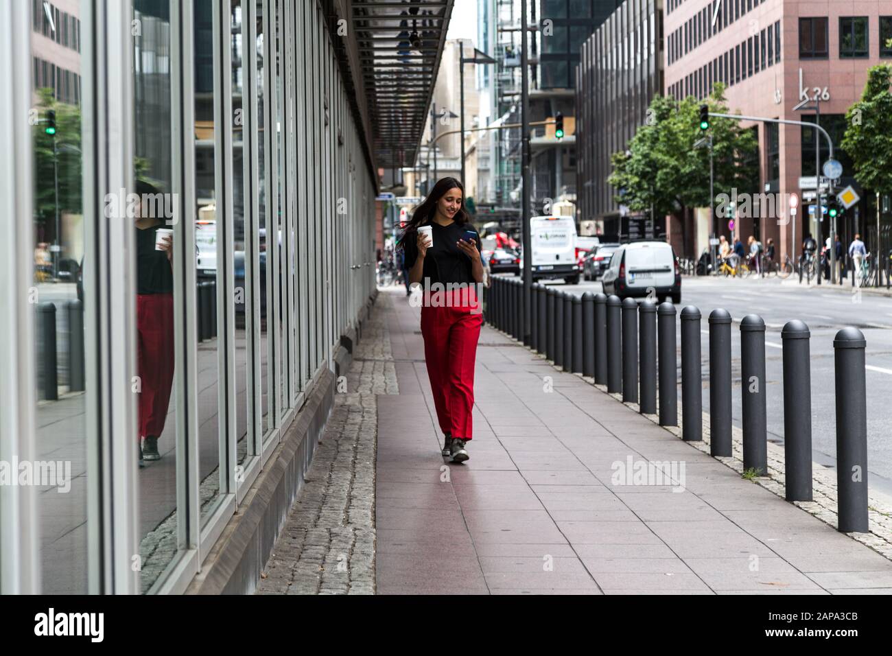 La jeune femme est heureuse de marcher sur le trottoir en tenant une tasse de café lors de ses déplacements et sur son téléphone portable. Tir long. Banque D'Images