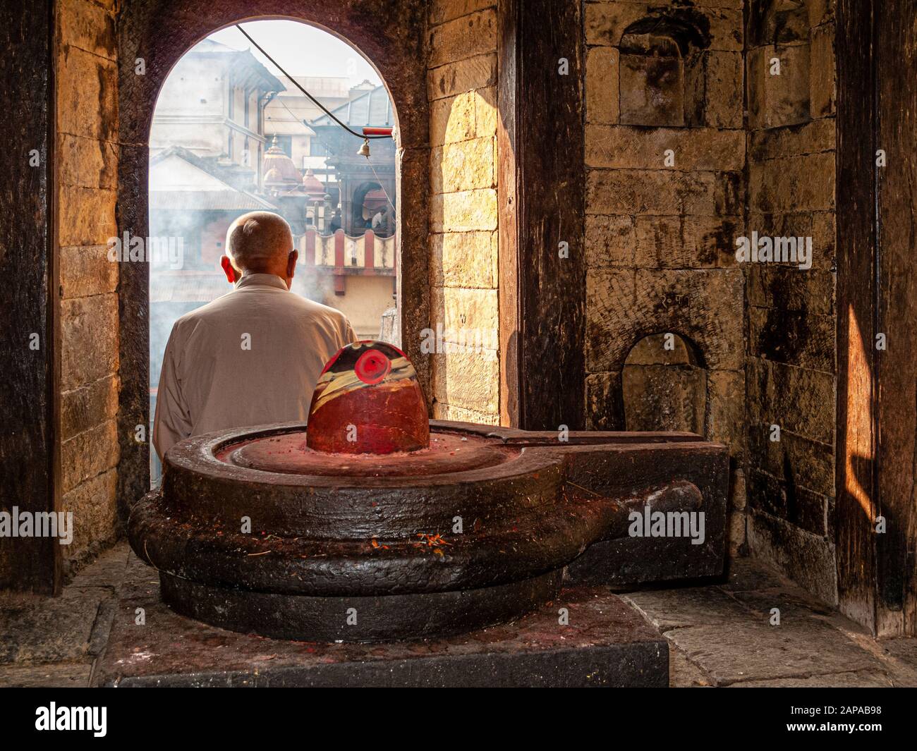 Prêtre local assis derrière un lingam Shiva dans un petit temple, en regardant une cérémonie de crémation au temple de Pashupatinath Banque D'Images