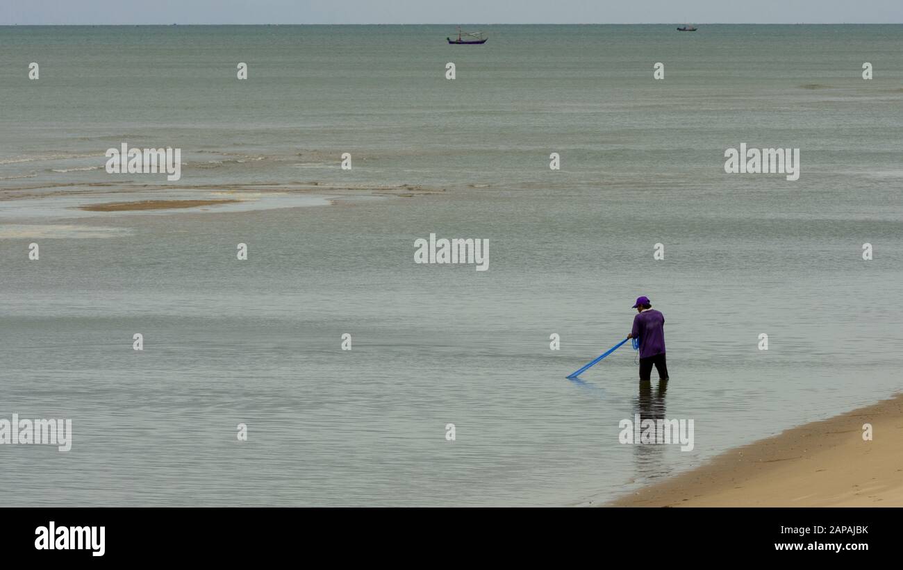 pêcheur thaïlandais avec chapeau bleu et chemise dans l'eau avec filet bleu à marée basse Banque D'Images