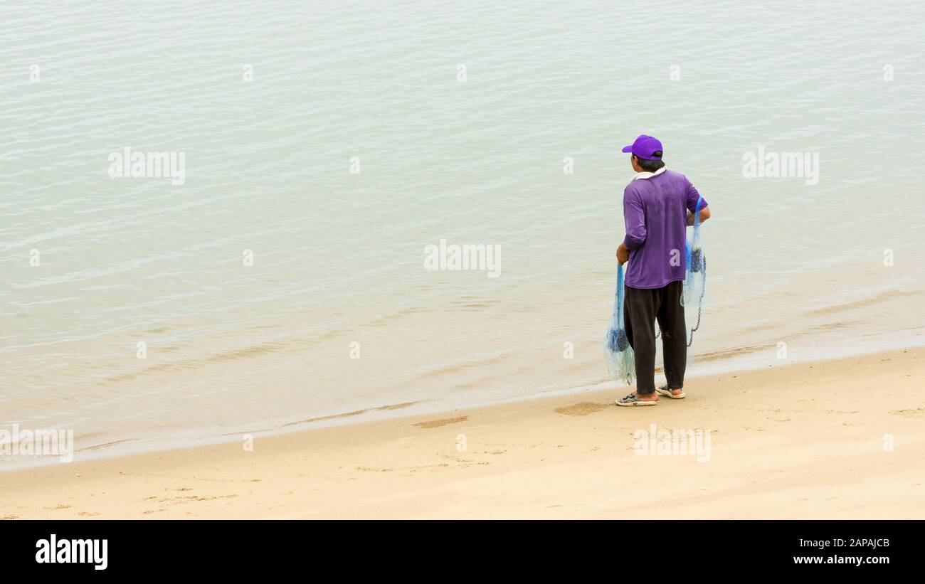 Pêcheur thaïlandais au bord de l'eau avec filet bleu à marée basse portant un chapeau violet et une chemise avec filet bleu Banque D'Images
