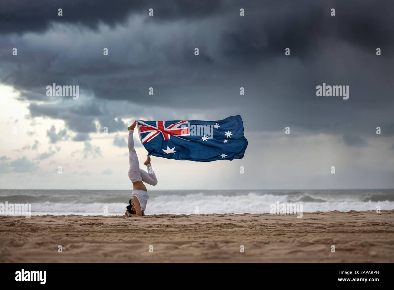 Drapeau australien qui souffle dans le vent suspendu par les pieds de la femelle yogi faisant une posture de tête sur une plage Aussie, avec des nuages de tempête dramatique fond Banque D'Images