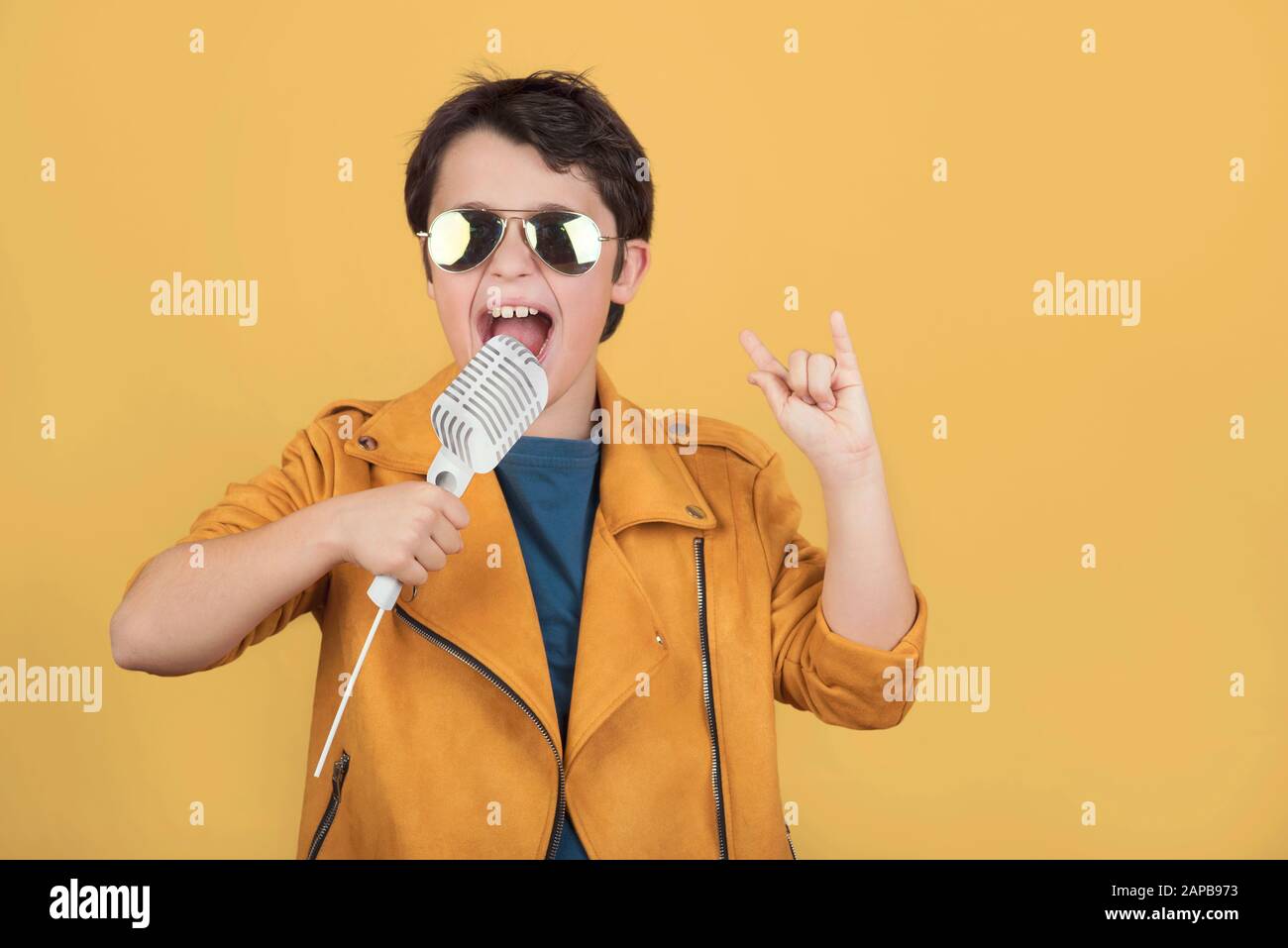 enfant avec lunettes de soleil tenant un microphone faisant le symbole rock avec les mains vers le haut sur fond jaune Banque D'Images