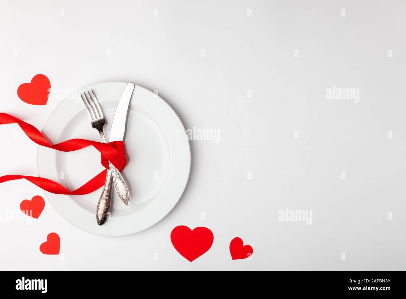 Table romantique sur fond blanc. Modèle de carte de Saint-Valentin. Ruban rouge, plaque, argenterie, fourche vintage, coeurs, couteau. Annonces de concept Banque D'Images