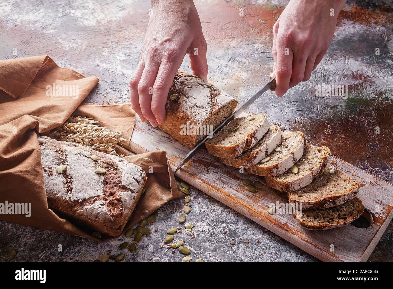 Les mains femelles coupent du pain de levain fraîchement cuit avec des graines de tournesol et de citrouille sur une serviette brune. Oreilles de blé. Prise de vue horizontale Banque D'Images