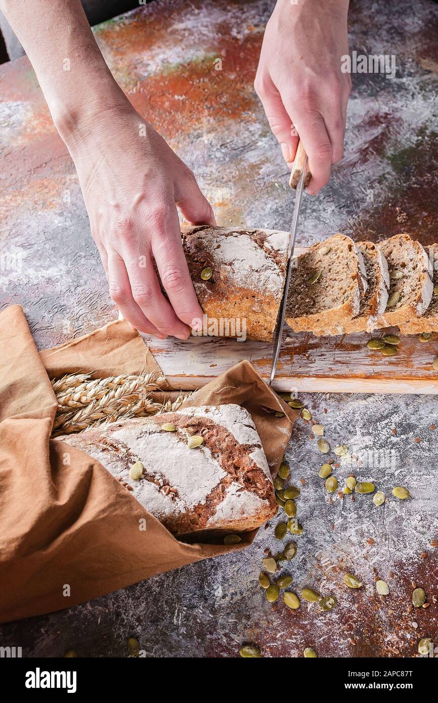 Les mains femelles à photo verticale coupent du pain de levain fraîchement cuit avec des graines de tournesol et de citrouille sur une serviette brune. Oreilles de blé. Banque D'Images