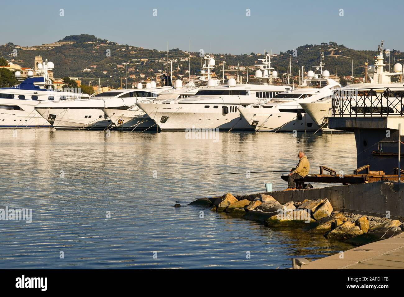 Pêcheur senior pêchant avec une tige dans le port de Porto Maurizio avec un yacht de luxe et la côte de la Riviera des fleurs en arrière-plan, Imperia Banque D'Images