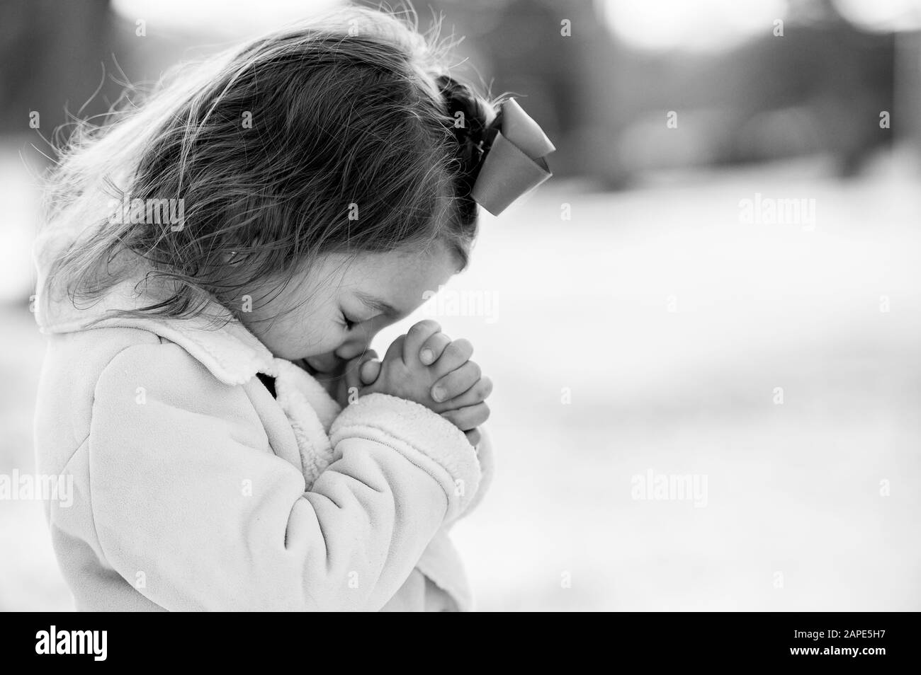 Échelle de gris d'une petite fille priant sous la lumière du soleil avec un arrière-plan flou Banque D'Images