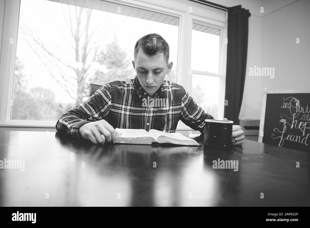 Échelle de gris d'un homme lisant un livre avec une tasse sur la barre de coupe sous les feux Banque D'Images