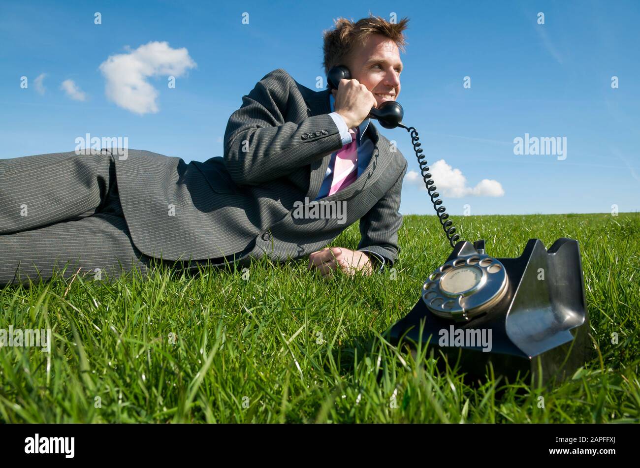 Un homme d'affaires gai se reposant sur un espace ensoleillé d'herbe verte vive, en parlant sur un téléphone à cadran rotatif à l'ancienne Banque D'Images