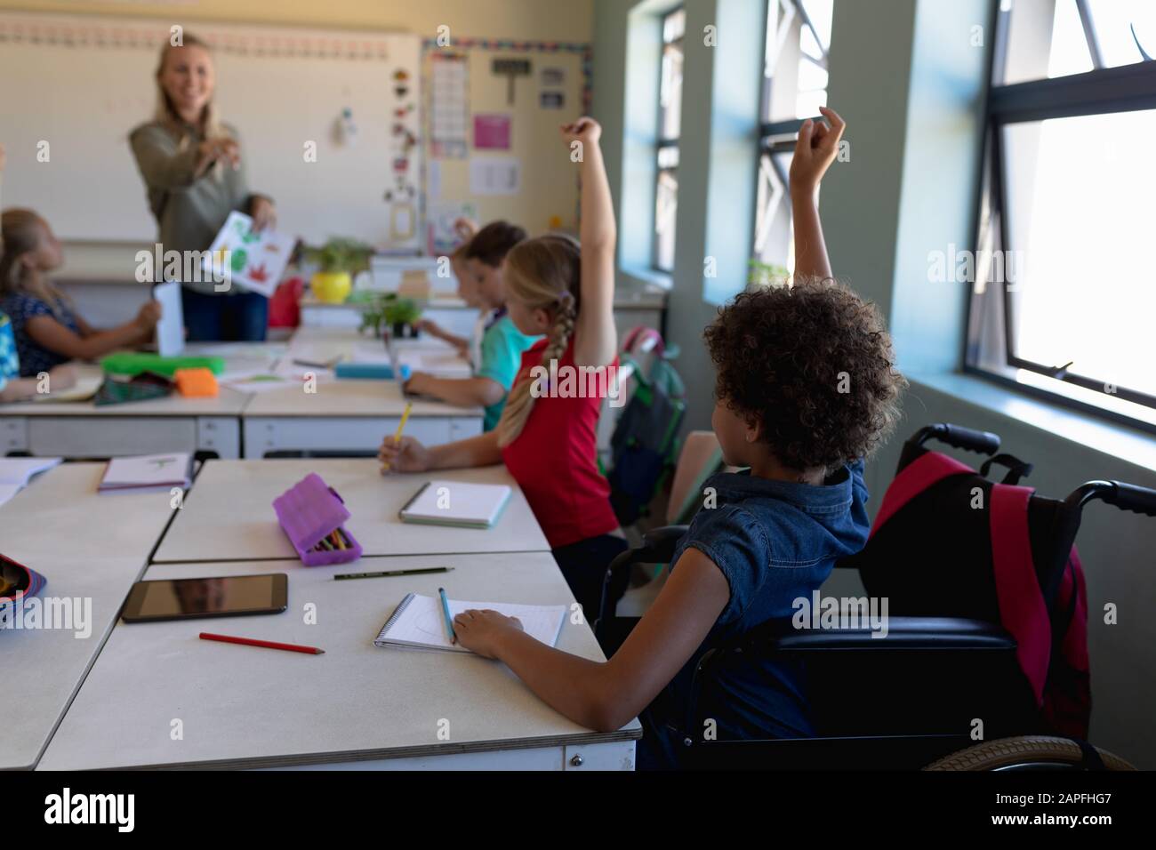 Groupe d'écoliers assis à un bureau levant les mains dans une classe d'école primaire Banque D'Images