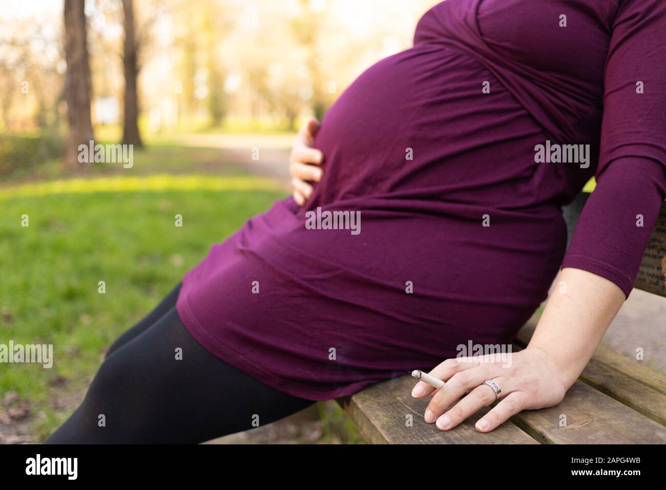 Gros plan sur la femme enceinte qui fume la cigarette dans sa main, assise sur le banc dans le parc. Faible profondeur de champ avec foyer sur la cigarette. Banque D'Images