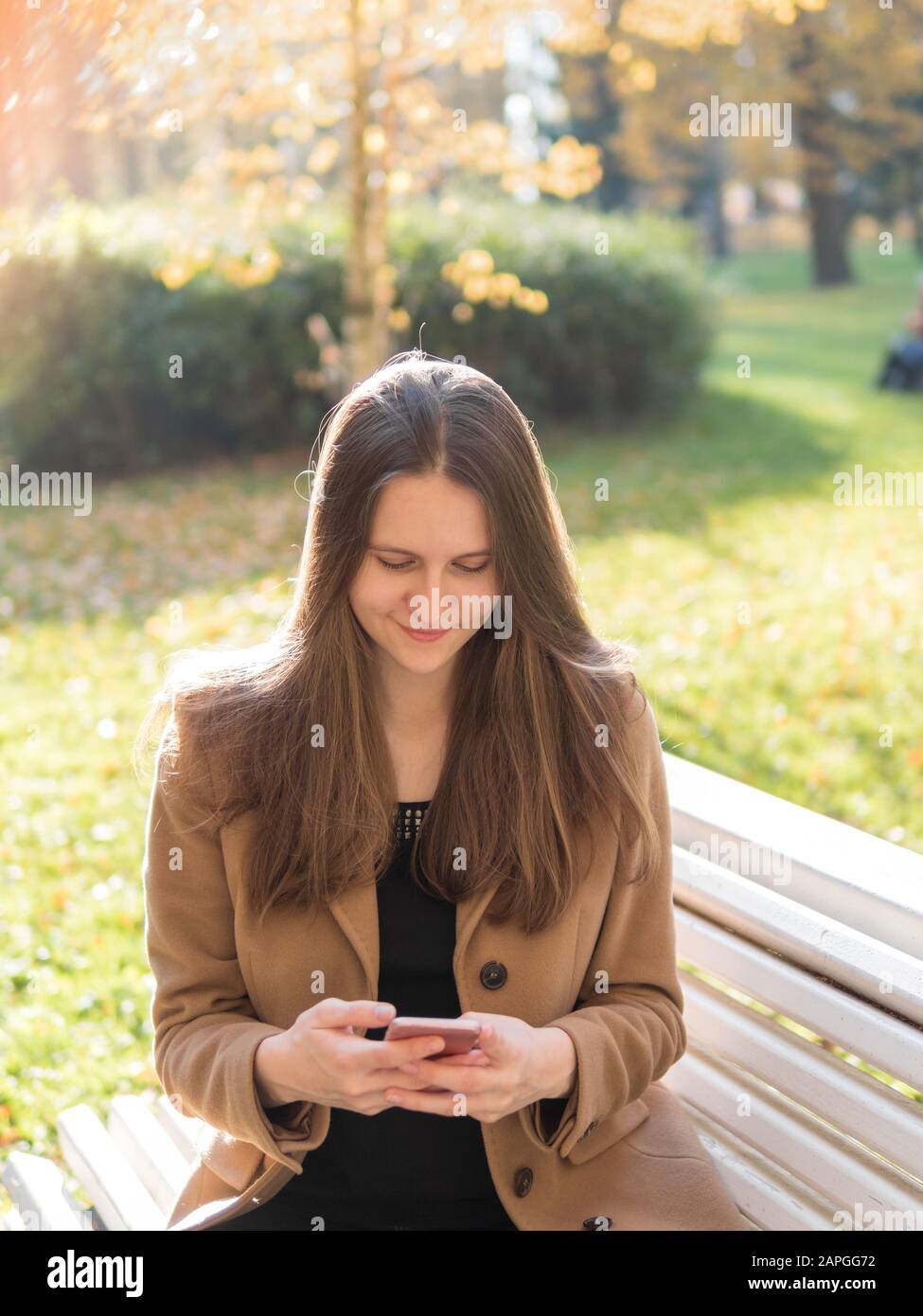 Belle jeune fille assise dans le parc sur un banc, tenant un smartphone et bavardant en ligne sur l'Internet. Femme aux cheveux longs Banque D'Images