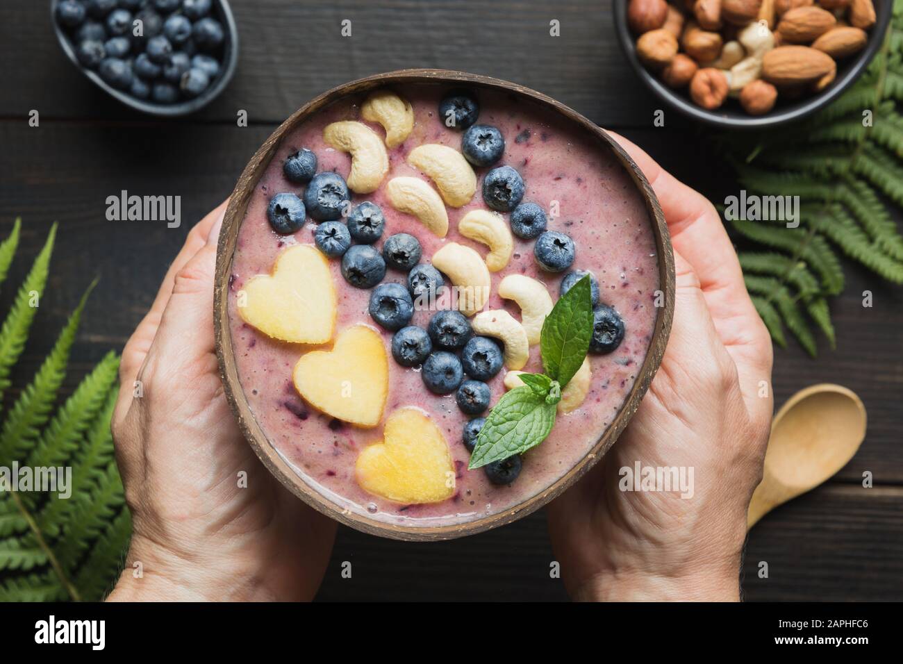 Smoothie tropicale à la banane et à la myrtille entre les mains des femmes. Petit déjeuner sain dans un bol à noix de coco. Vue de dessus. Manger propre, cuisine végétarienne. Banque D'Images