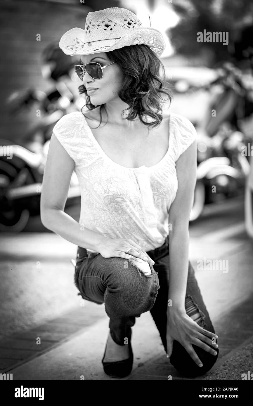 Image en noir et blanc de la femme africaine américaine avec lunettes de soleil et chapeau de cow-boy en paille blanche s'agenouillant sur le sol avec moto de rue et wal en brique rouge Banque D'Images