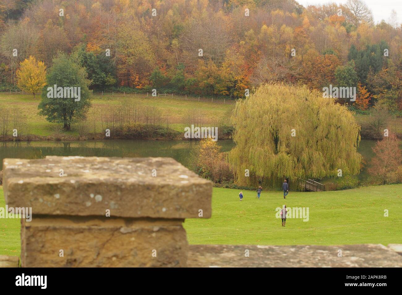 Vue sur un mur de jardin en pierre à l'herbe en pente avec une jeune famille jusqu'à un lac avec des arbres d'automne et un bois au-delà Banque D'Images