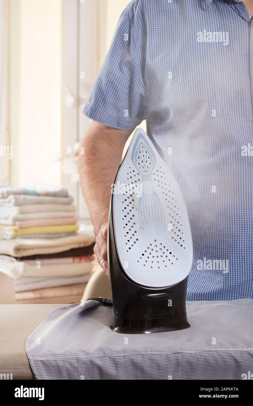 Le fer noir se tient sur le pied de la planche à repasser. La vapeur s'échappe de la partie argentée. Un homme repassage des vêtements. En arrière-plan, repassé et folie Banque D'Images