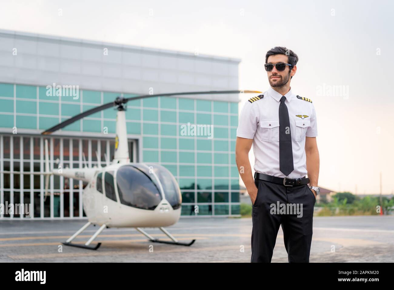 Portrait de beau pilote commercial en uniforme blanc capitaine debout et regardant intelligent près d'un petit hélicoptère privé sur un point d'atterrissage avec privé Banque D'Images