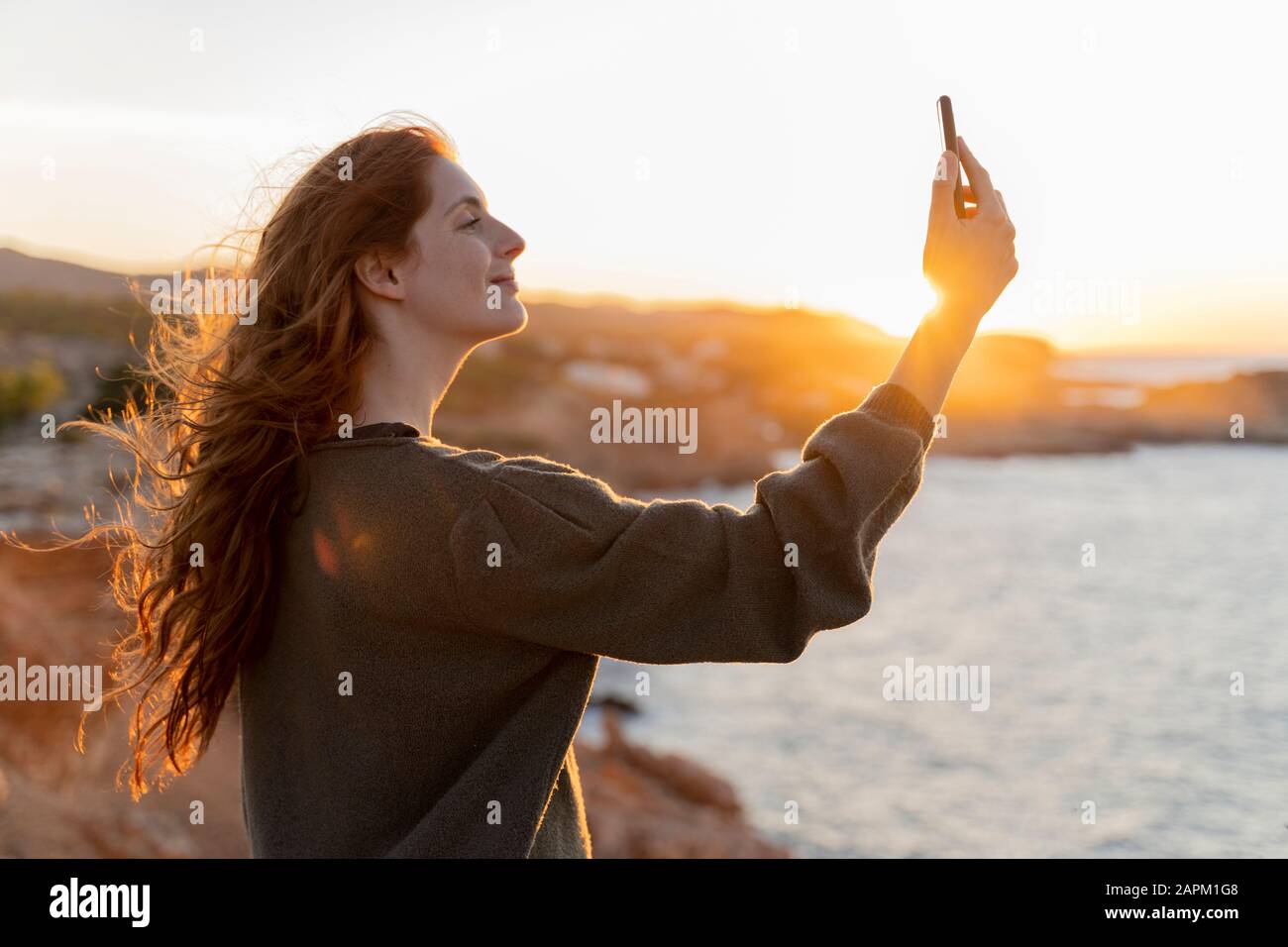 Jeune femme à tête rouge prenant un selfie sur la côte au coucher du soleil, Ibiza, Espagne Banque D'Images