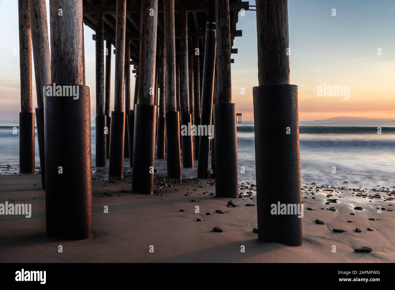 Gros plan des piliers, Ventura Pier, Ventura, Californie au coucher du soleil. Sable et rochers en premier plan; océan soyeux, ciel coloré au-delà. Banque D'Images