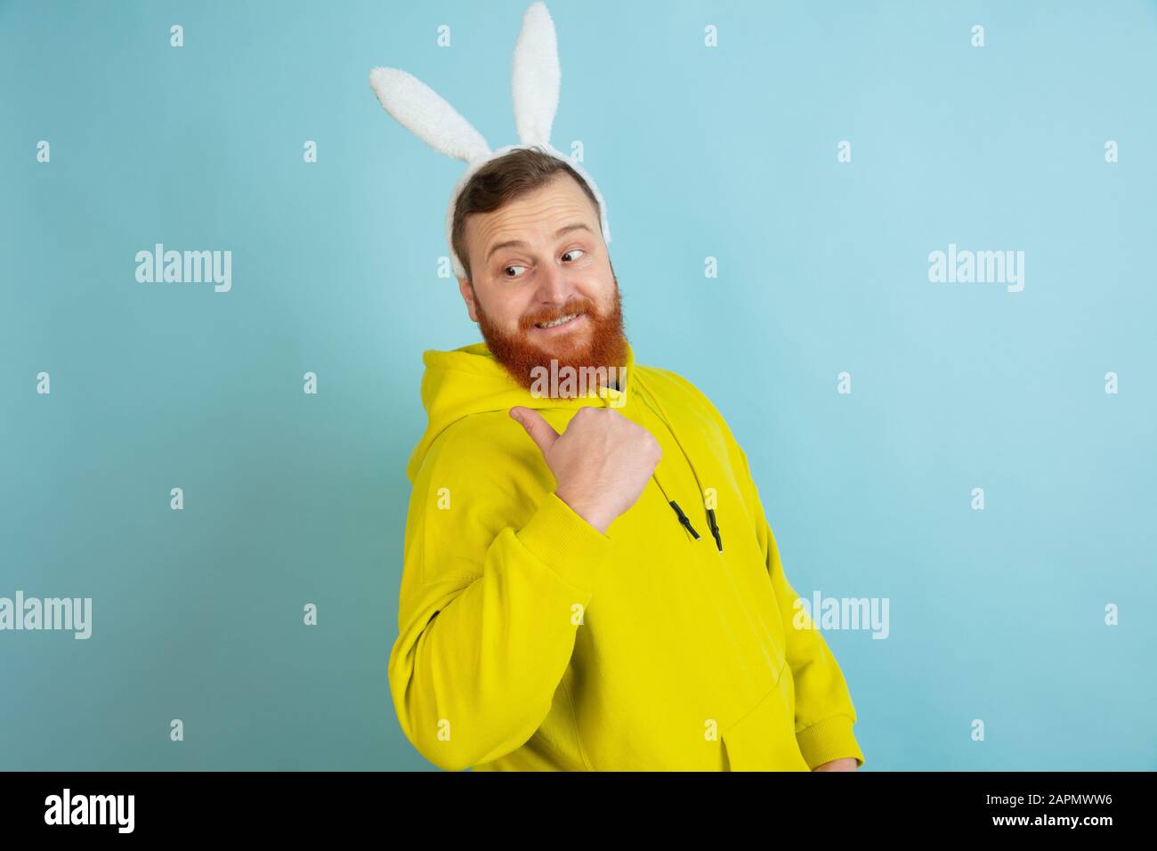 Pointant vers l'arrière, souriant. Homme caucasien comme lapin de Pâques avec des vêtements décontractés lumineux sur fond bleu studio. Joyeuses salutations de pâques. Concept d'émotions humaines, d'expression faciale, de vacances. Copyspace. Banque D'Images