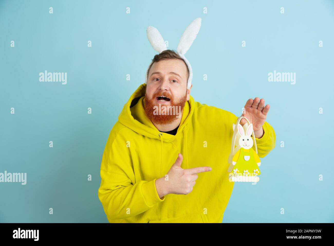 Pointant sur la décoration. Homme caucasien comme lapin de Pâques avec des vêtements décontractés lumineux sur fond bleu studio. Joyeuses salutations de pâques. Concept d'émotions humaines, d'expression faciale, de vacances. Copyspace. Banque D'Images
