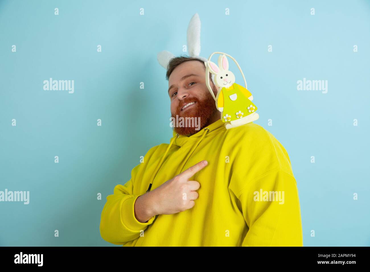 Pointant sur la décoration. Homme caucasien comme lapin de Pâques avec des vêtements décontractés lumineux sur fond bleu studio. Joyeuses salutations de pâques. Concept d'émotions humaines, d'expression faciale, de vacances. Copyspace. Banque D'Images