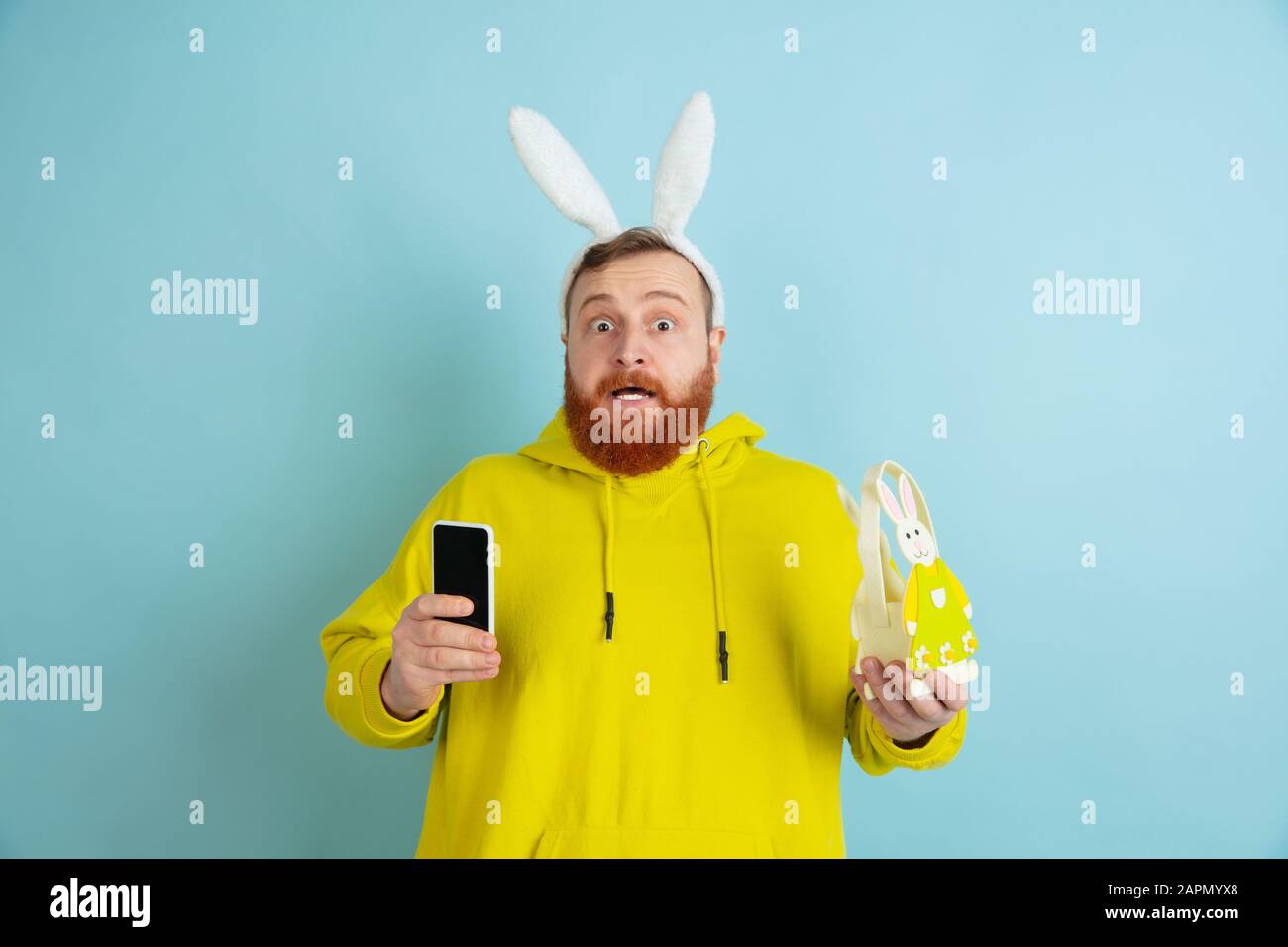 Homme caucasien comme lapin de Pâques avec des vêtements et un décor clairs et décontractés prenant le selfie sur fond bleu studio. Joyeuses salutations de pâques. Concept d'émotions humaines, d'expression faciale, de vacances. Copyspace. Banque D'Images