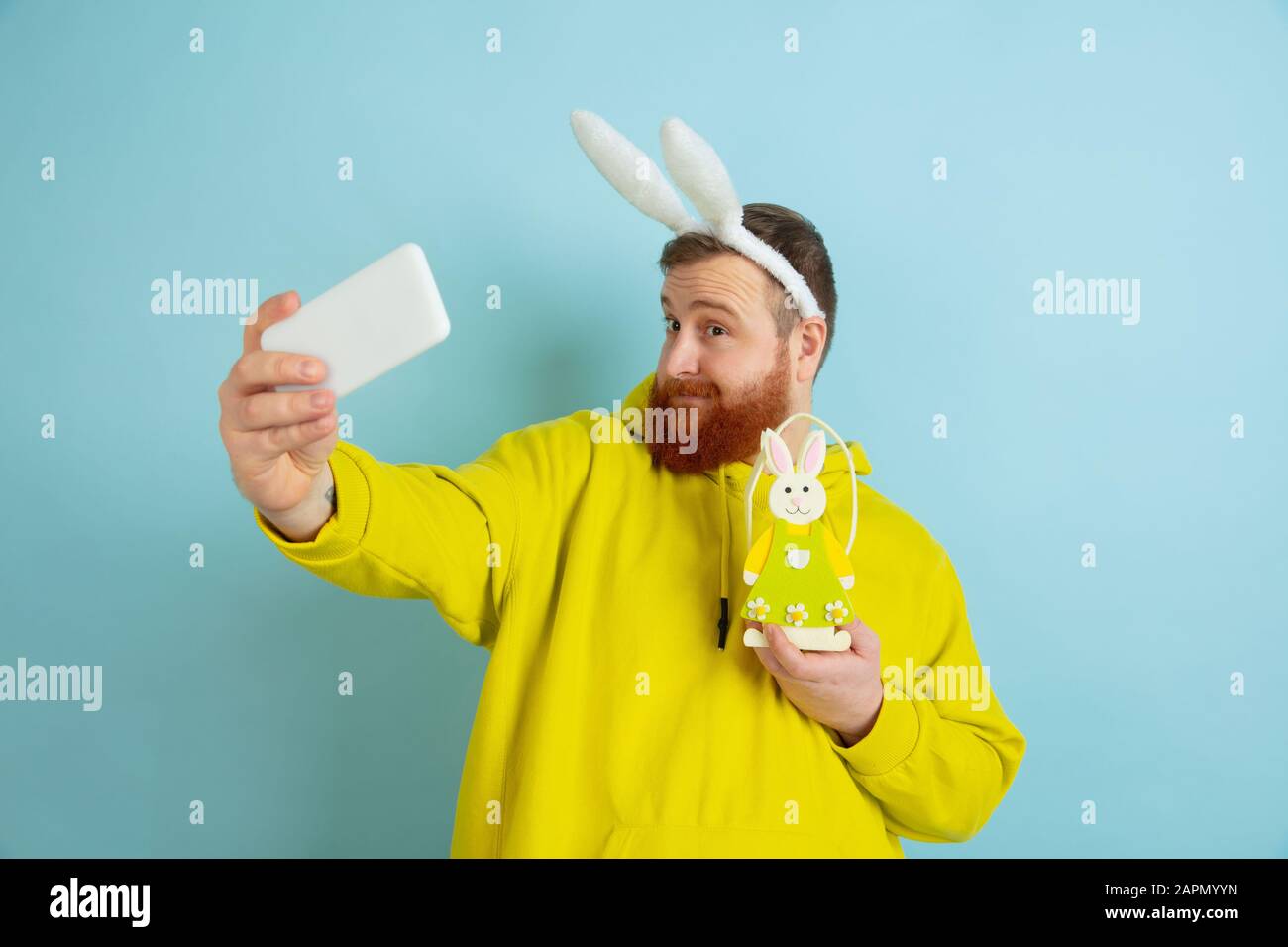 Homme caucasien comme lapin de Pâques avec des vêtements et un décor clairs et décontractés prenant le selfie sur fond bleu studio. Joyeuses salutations de pâques. Concept d'émotions humaines, d'expression faciale, de vacances. Copyspace. Banque D'Images