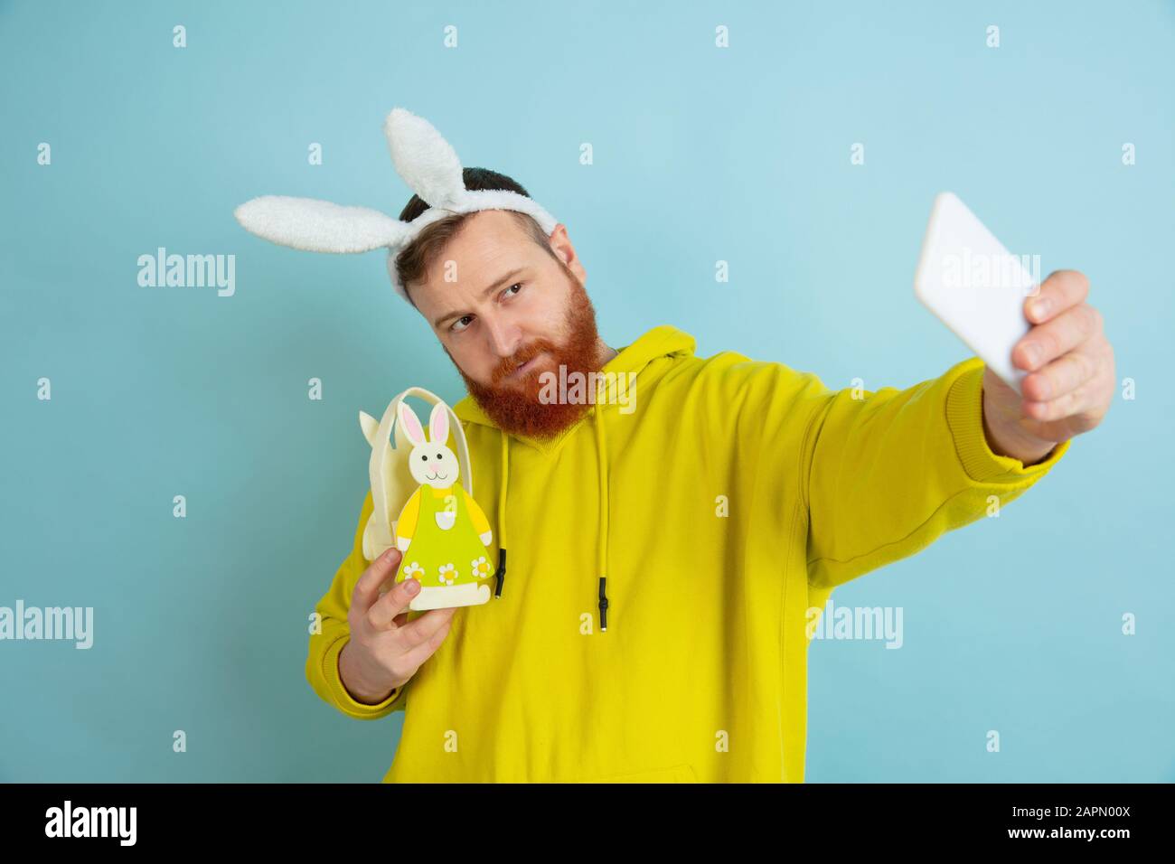 Homme caucasien comme lapin de Pâques avec des vêtements et un décor clairs et décontractés prenant le selfie sur fond bleu studio. Joyeuses salutations de pâques. Concept d'émotions humaines, d'expression faciale, de vacances. Copyspace. Banque D'Images