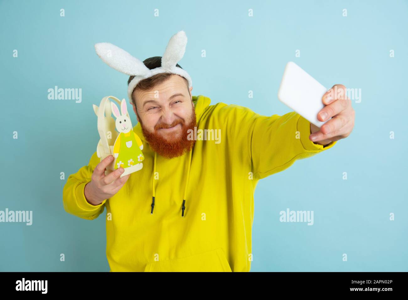 Homme caucasien comme lapin de Pâques avec des vêtements et un décor clairs et décontractés prenant le selfie sur fond bleu studio. Joyeuses salutations de pâques. Concept d'émotions humaines, d'expression faciale, de vacances. Copyspace. Banque D'Images