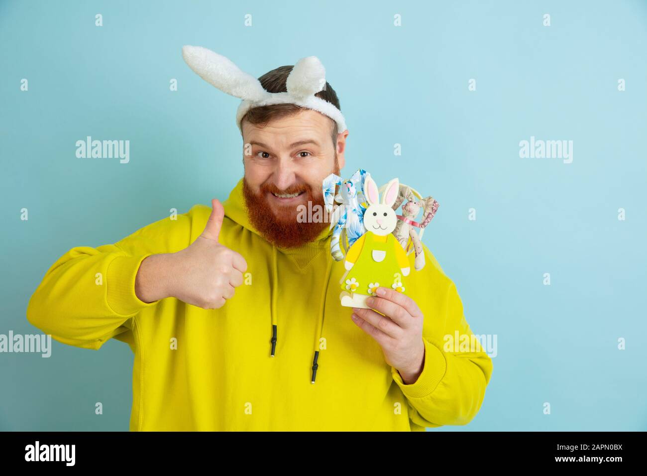 Avec le pouce vers le haut. Homme caucasien comme lapin de Pâques avec des vêtements décontractés lumineux sur fond bleu studio. Joyeuses salutations de pâques. Concept d'émotions humaines, d'expression faciale, de vacances. Copyspace. Banque D'Images