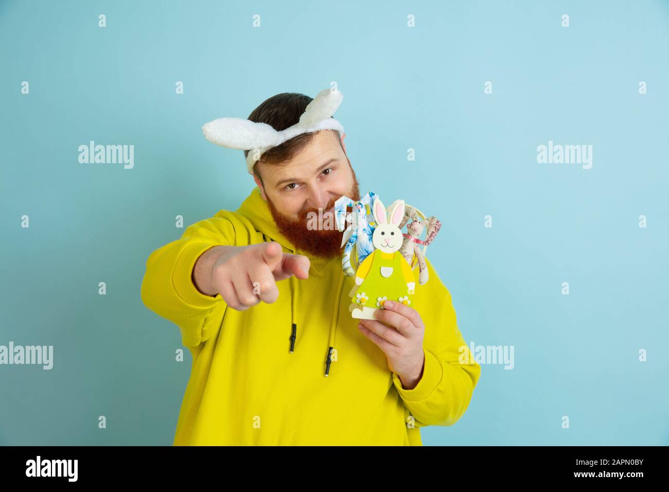 Pointant, vous choisir. Homme caucasien comme lapin de Pâques avec des vêtements décontractés lumineux sur fond bleu studio. Joyeuses salutations de pâques. Concept d'émotions humaines, d'expression faciale, de vacances. Copyspace. Banque D'Images
