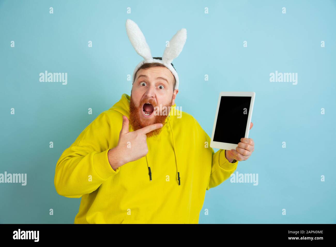 Pointant sur l'écran de la tablette. Homme caucasien comme lapin de Pâques avec des vêtements décontractés et lumineux sur fond bleu. Joyeuses salutations de pâques. Concept d'émotions humaines, d'expression faciale, de vacances. Copyspace. Banque D'Images