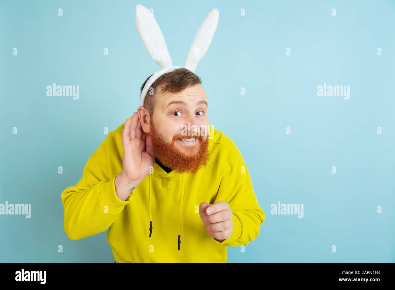 Ecouter le secret. Homme caucasien comme lapin de Pâques avec des vêtements décontractés lumineux sur fond bleu studio. Joyeuses salutations de pâques. Concept d'émotions humaines, d'expression faciale, de vacances. Copyspace. Banque D'Images