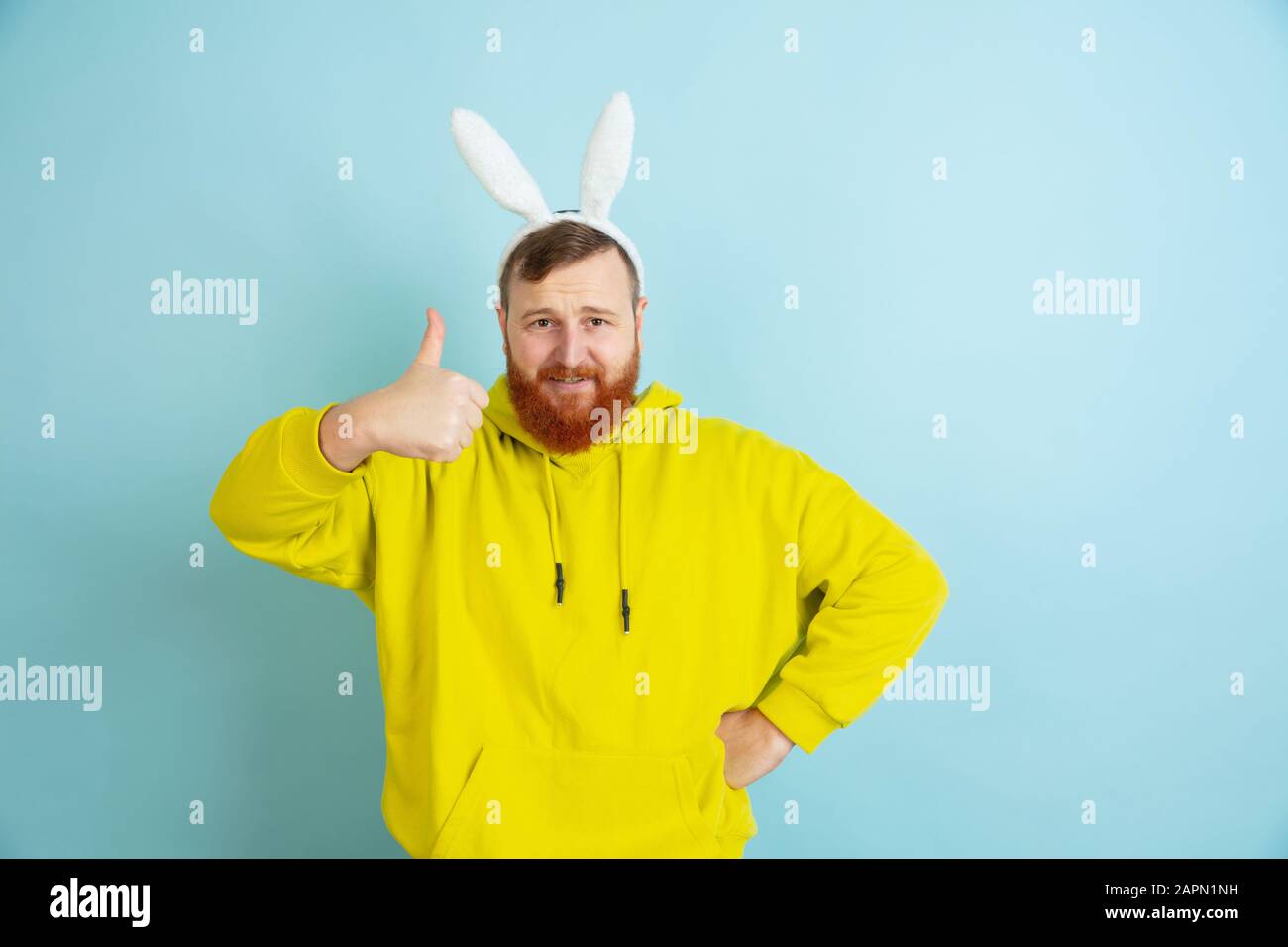 Avec le pouce vers le haut. Homme caucasien comme lapin de Pâques avec des vêtements décontractés lumineux sur fond bleu studio. Joyeuses salutations de pâques. Concept d'émotions humaines, d'expression faciale, de vacances. Copyspace. Banque D'Images
