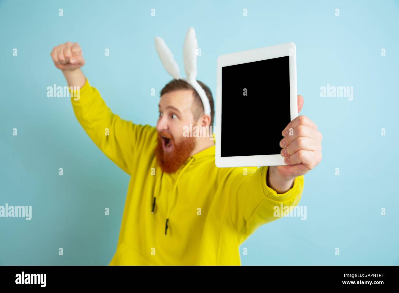 Affichage de l'écran de la tablette. Homme caucasien comme lapin de Pâques avec des vêtements décontractés lumineux sur fond bleu studio. Joyeuses salutations de pâques. Concept d'émotions humaines, d'expression faciale, de vacances. Copyspace. Banque D'Images