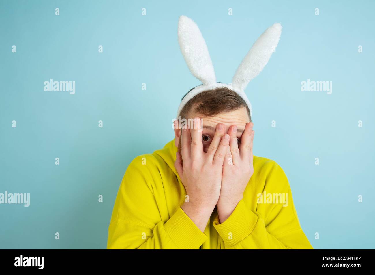 Peur, se cacher le visage dans les mains. Homme caucasien comme lapin de Pâques avec des vêtements décontractés et lumineux sur fond bleu. Joyeuses salutations de pâques. Concept d'émotions humaines, d'expression faciale, de vacances. Copyspace. Banque D'Images