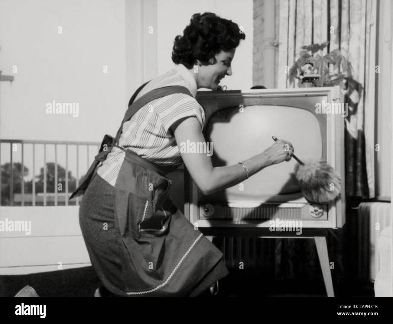 Une femme au foyer avec un tablier dépoussiérant un téléviseur avec un dépoussiéreur. Banque D'Images Une femme au foyer avec un tablier dépoussiérant un téléviseur avec un dépoussiéreur. Banque D'Images