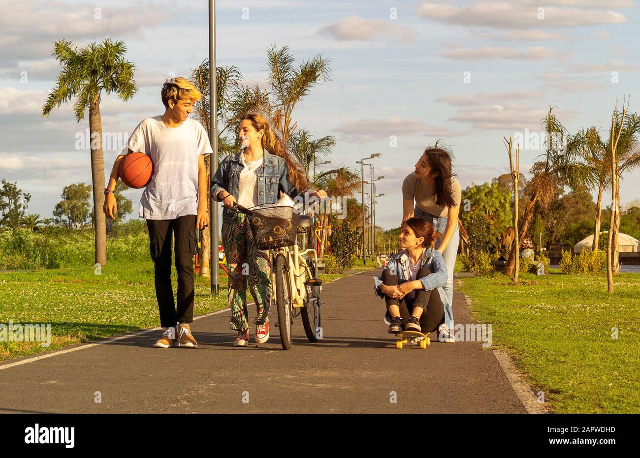 Portrait avant de quatre adolescents marchant le long d'un sentier dans un parc avec un vélo, un skateboard, des patins à roulettes et un basket-ball Banque D'Images