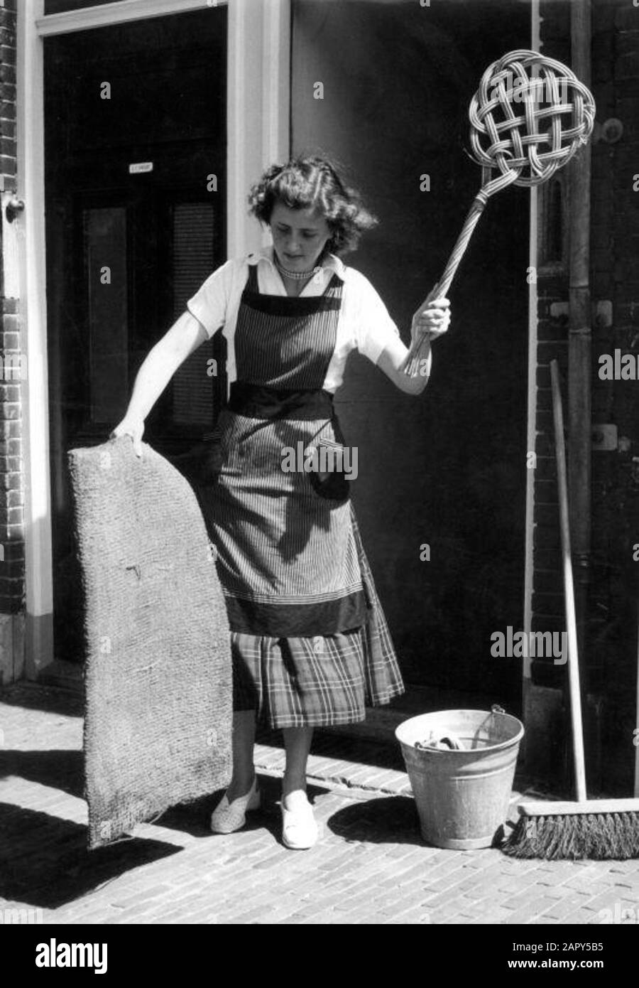 Tapis de sol. La femme de ménage néerlandaise avec tablier frappe le tapis  avec un défonce de tapis, dans la rue près de la porte d'entrée. À côté  d'elle un balai et