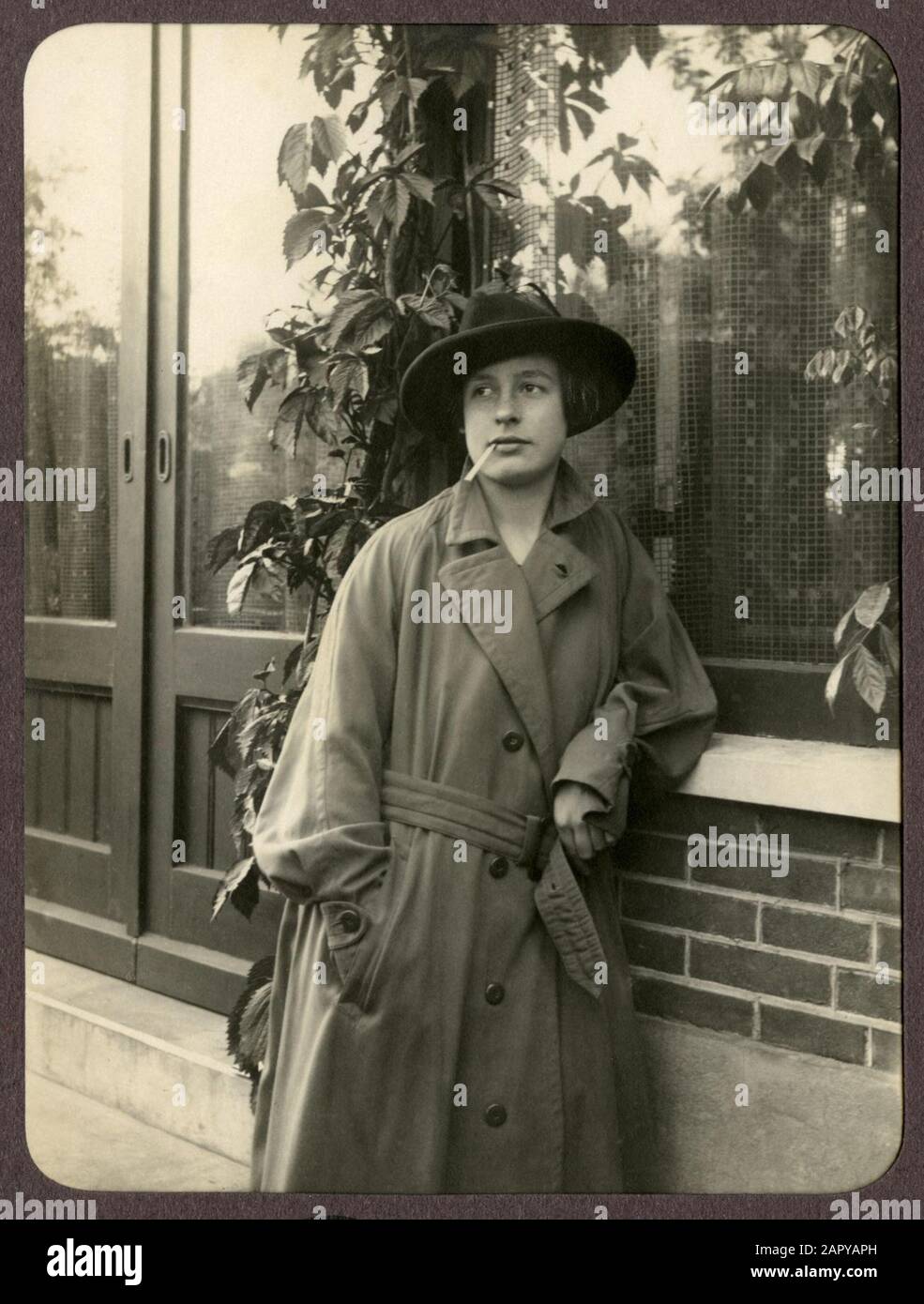 Femme en long imperméable avec chapeau et cigarette dans la bouche, penchée contre une fenêtre. Pays-Bas, 1926. Cette photo fait partie d'un certain nombre de photos numérisées en basse résolution d'un toalbum privé. Banque D'Images